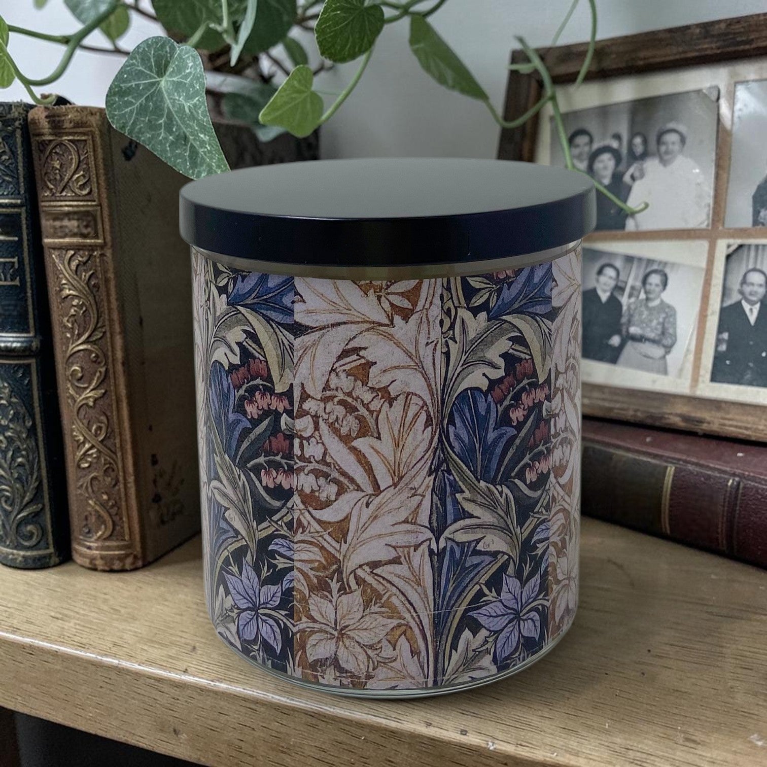 William Morris artwork design candle with floral and acanthus pattern on a wooden surface with books and family photos in the background.