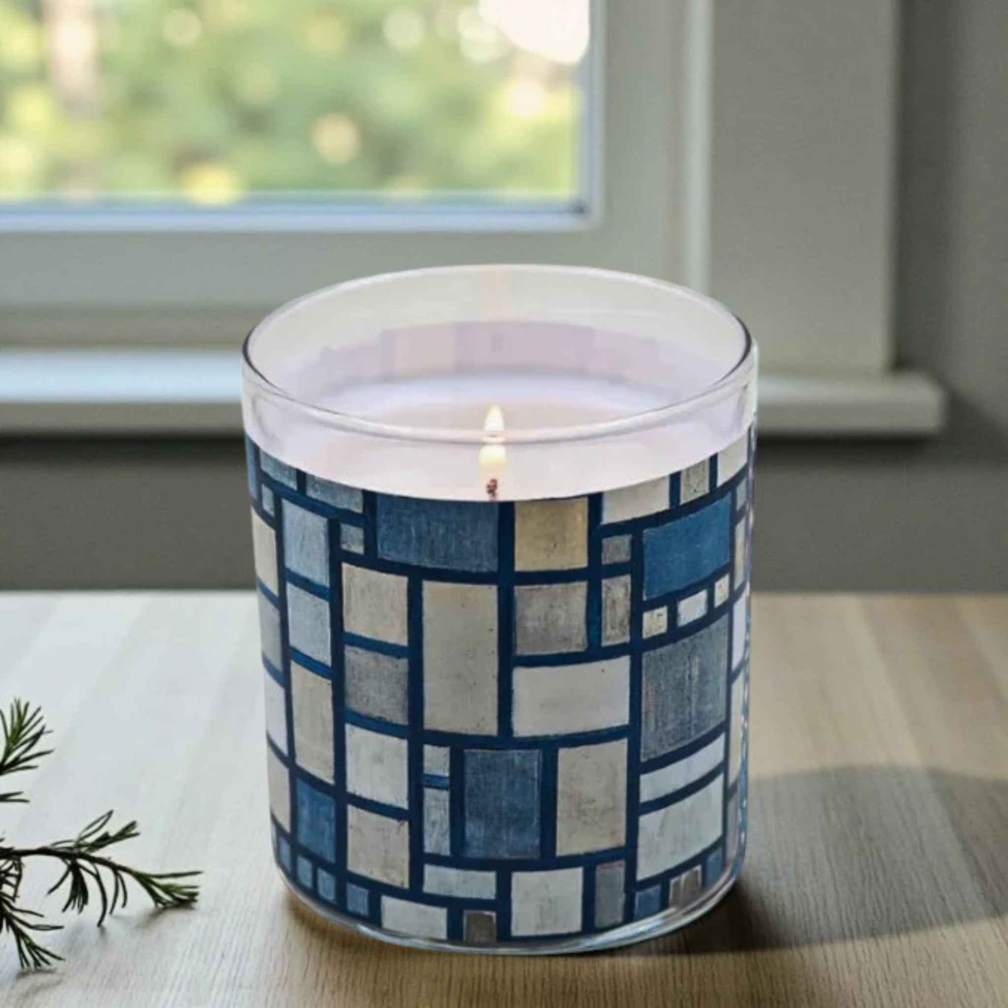 Candle in a glass jar with a blue geometric pattern on a table near a window.