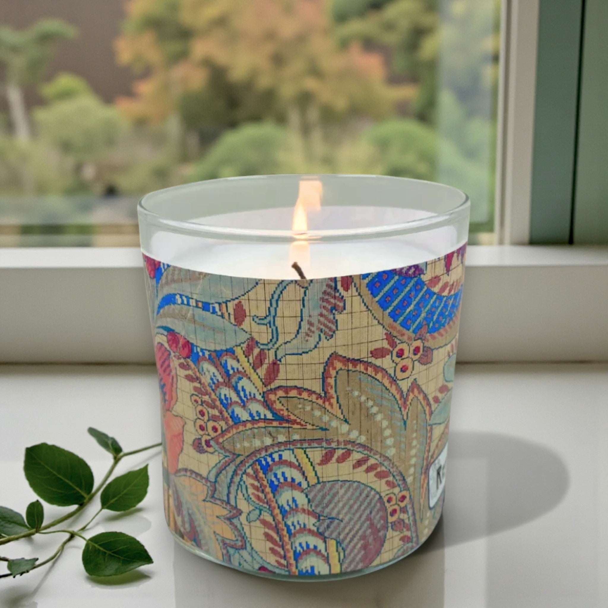 Candle with a colorful patterned label on a windowsill with greenery outside.