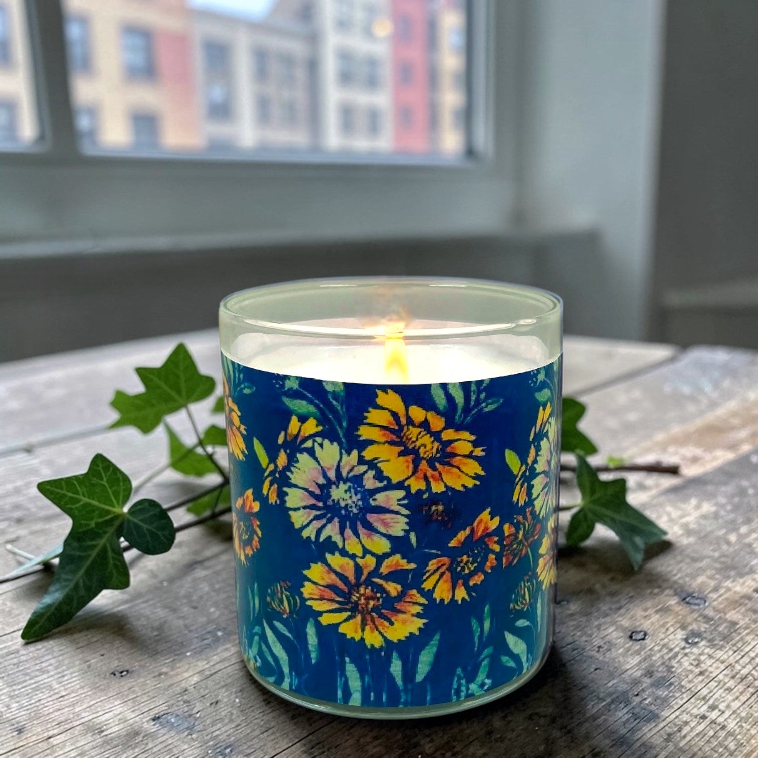 Candle with a floral design on a blue label, placed on a wooden surface with green leaves.