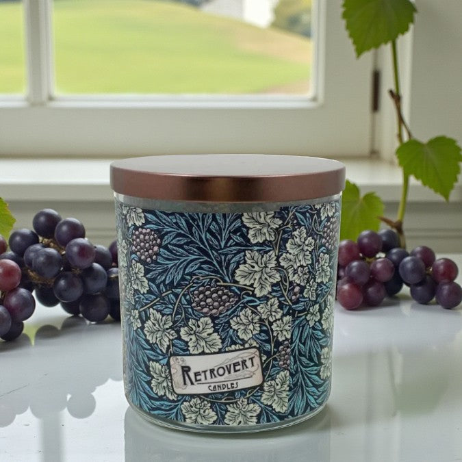 Decorative candle with floral design and wooden lid on a surface with grapes and a window in the background