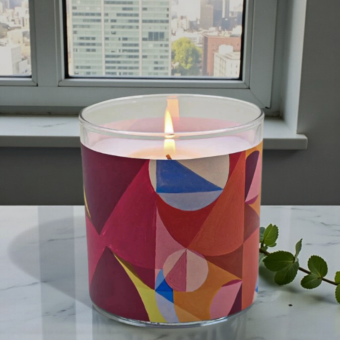 Candle with a colorful geometric design in a clear container, burning on a marble surface with a cityscape in the background.