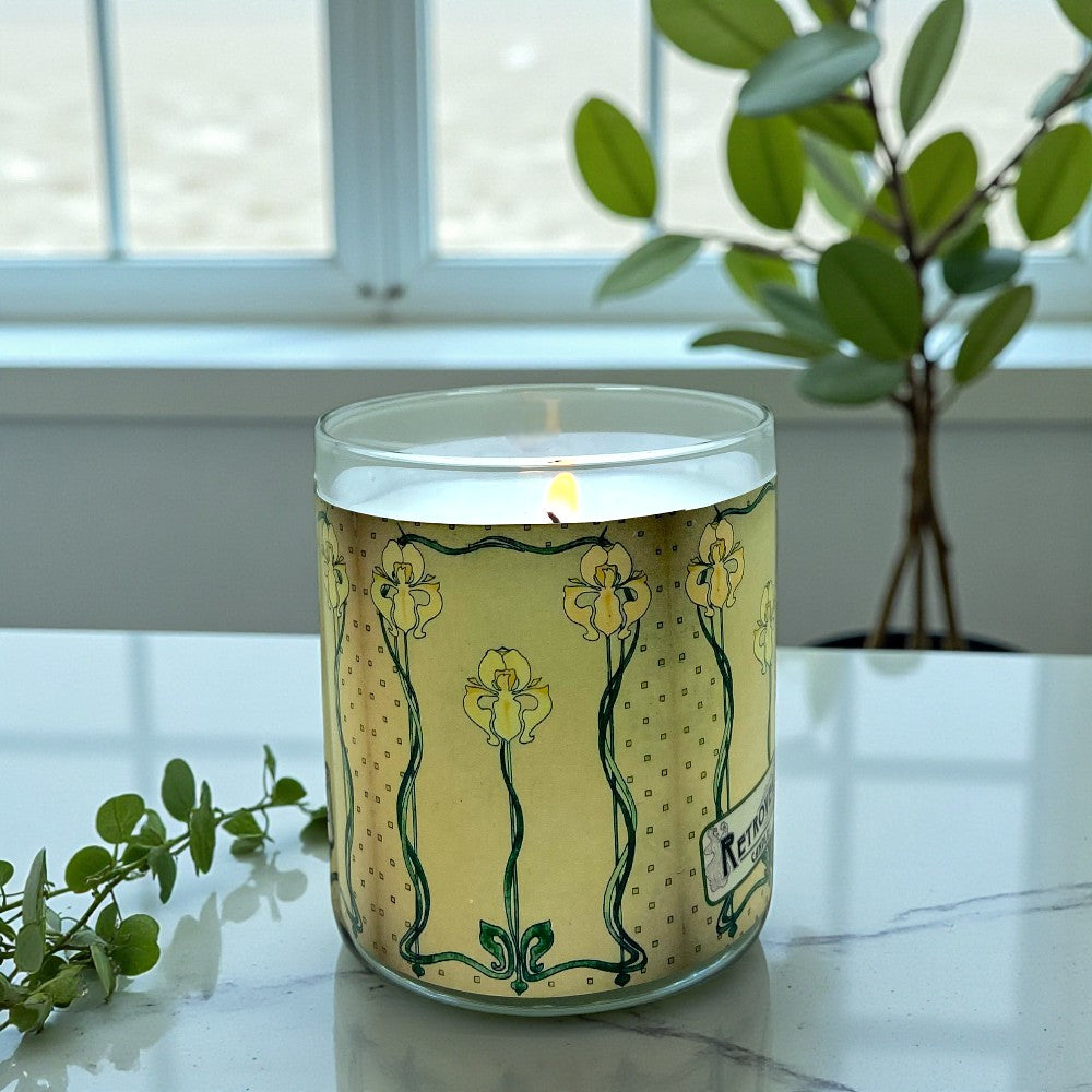 Candle in a glass jar with a decorative label on a marble surface, surrounded by greenery.