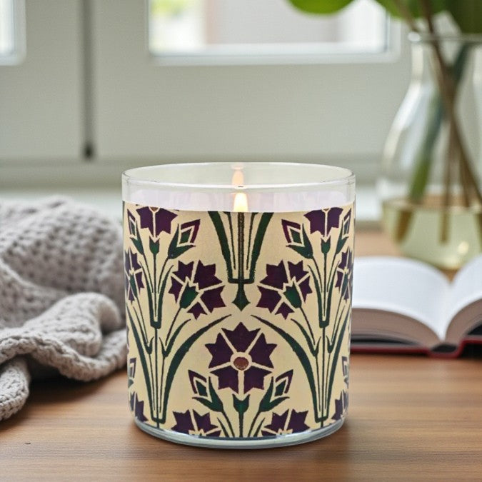 Candle with floral pattern on a wooden surface, with a book and plant in the background.