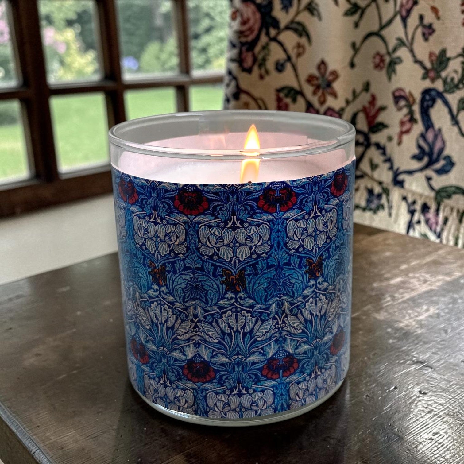 Candle in a decorative blue and red patterned container on a wooden surface with a floral curtain in the background.